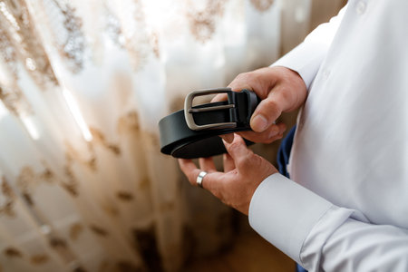Man preparing for an event holding a black leather belt, showing elegant and sophisticated style