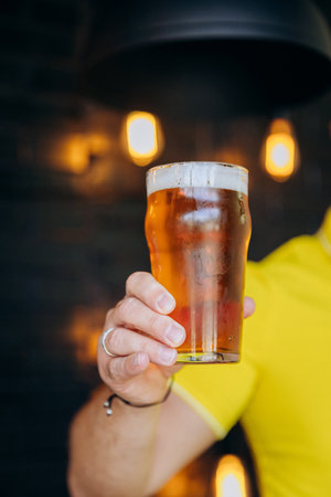 Close-up of person holding glass of beer with blurred pub background. Beer-related contentの写真素材