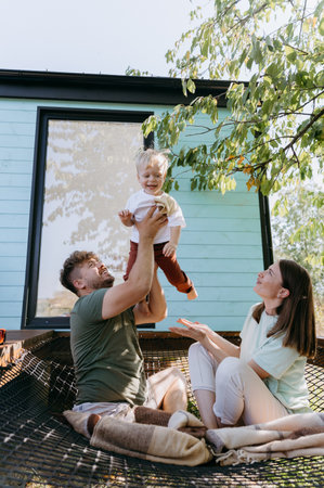 Father lifting his happy toddler in air while mom watches with joy, playful family moments outdoors