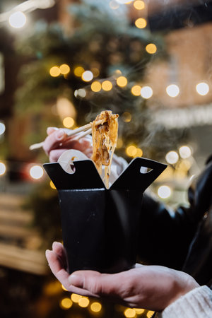 Steaming Asian noodles in a takeaway box under festive city lights, perfect for an evening snackの写真素材