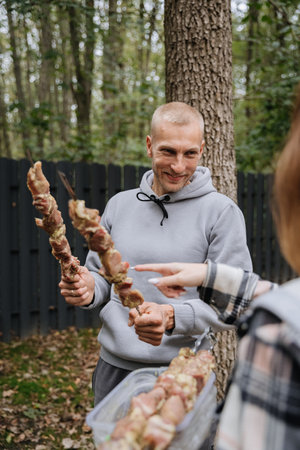 Man holding skewers with marinated meat, smiling while preparing for barbecue in cozy forest settingの写真素材