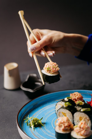 Hand using chopsticks to hold sushi roll with crab topping, served on a blue plate with soy sauceの写真素材