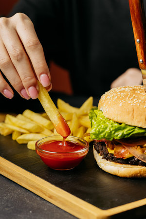 Dipping crispy French fry into ketchup beside a juicy burger with fresh lettuce, food momentの写真素材