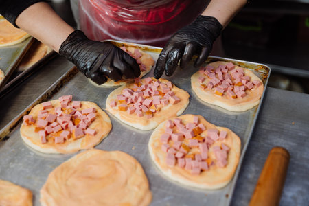 Preparing mini pizzas with diced ham and sauce, perfect for baking in a commercial kitchen