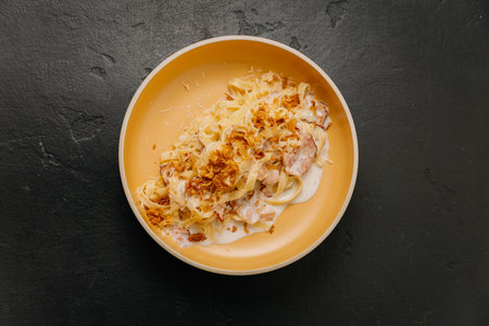 Creamy tagliatelle pasta with crispy bacon and fried onions in a beige bowl on a dark backgroundの写真素材