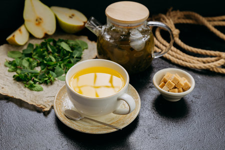 Pear and mint herbal tea in cup with glass teapot, fresh ingredients, and sugar cubes on dark tableの写真素材