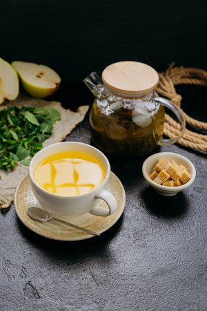 Pear and mint herbal tea in cup with glass teapot, fresh ingredients, and sugar cubes on dark tableの写真素材