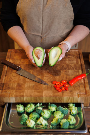 Hands holding fresh halved avocado on wooden board with chili and broccoli