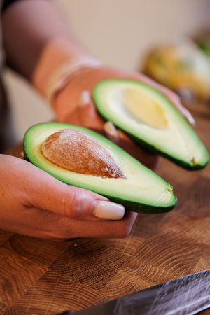 Freshly sliced avocado halves held in hands on wooden cutting board with vegetablesの写真素材