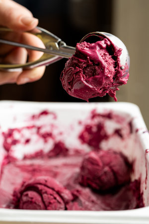 Hand holding scoop of berry sorbet over container, closeup view of vibrant frozen dessert