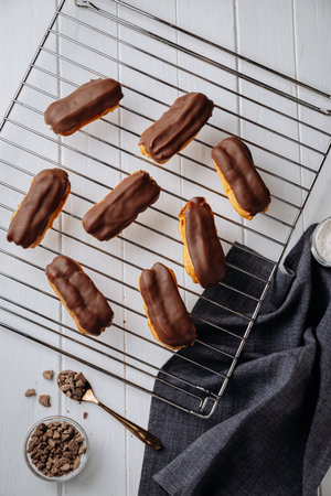 Mini chocolate eclairs on a cooling rack over white wooden table, flat lay view