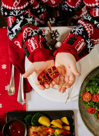 Child in festive sweater holding red gift boxes over Christmas dinner table celebrationの写真素材