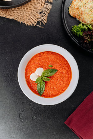 Tomato soup puree with basil and mozzarella in white bowl on dark tabletop, top view