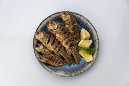 Crispy whole fried fish with lemon wedges and basil on ceramic plate, overhead on white