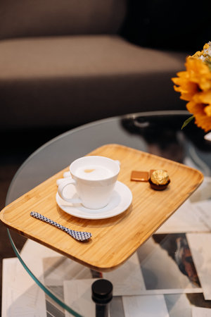 Cappuccino on wooden tray with chocolates in cozy lounge, glass table and flowersの写真素材