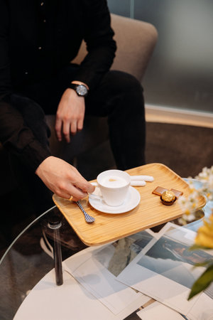 Man taking cappuccino from wooden tray with chocolates in lounge, glass table, close up