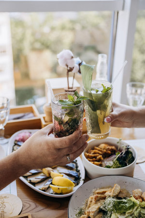 Couple clinking fresh summer cocktails at restaurant table, brunch toast by window, copy space