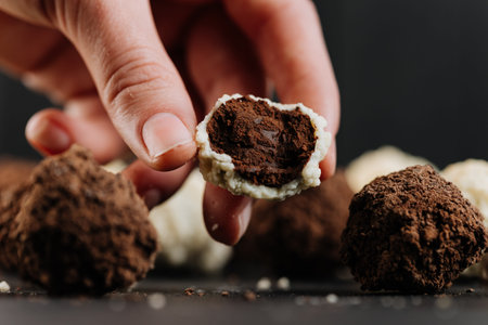 Hand holding cut coconut truffle with dark chocolate filling, macro on blackの写真素材