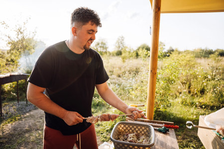 Man preparing meat skewers for outdoor barbecue at sunset, summer grilling in natureの写真素材