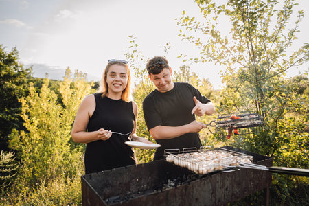Couple grilling vegetables outdoors at sunset, tasting food at summer barbecue