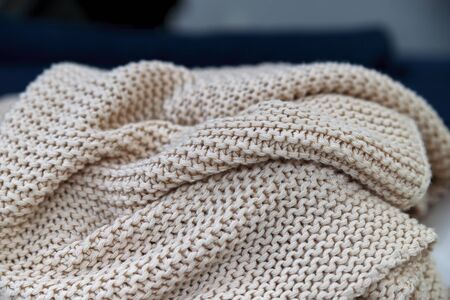 A soft knitted beige blanket made of natural wool lies rolled up against the background of a washed-out background. A close-up.の写真素材