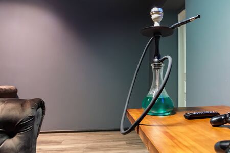 Black matte hookah with clear water with a black hose on the table next to the remote control in the...
