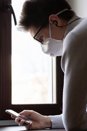 A young man in a reusable medical mask looks at the phone screen for information on how to protect...