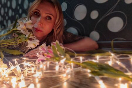 Portrait of a beautiful fashionable middle-aged woman with long blond hair, lying among white flowers and a garland of lights.の写真素材