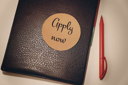 The message APPLY NOW on an orange sticker on a notepad lying on the desktopの写真素材