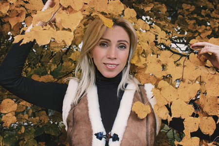 Blonde with long hair on a background of yellow leaves. Smiling woman in fashionable warm vest in retro style Woman on a walk in the autumn forest. healthy lifestyleの写真素材