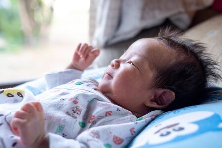 The adorable cute newborn baby infant with big black eyes lying on the bed with the view of garden on the window backgroundの写真素材