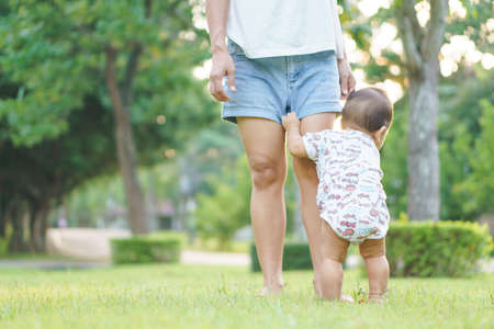 Mother helping Asian baby to learn first step on a lawn in summer day