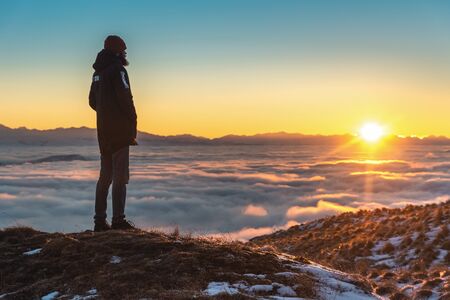 A man stands high in the mountains, at sunset, above the clouds.