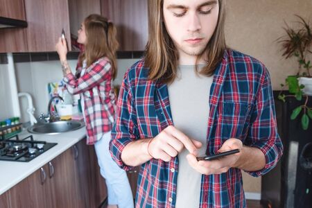 A young man talks on the phone in the kitchen at home while his wife is cleaning. The concept of family life, resolving disputes and problemsの写真素材