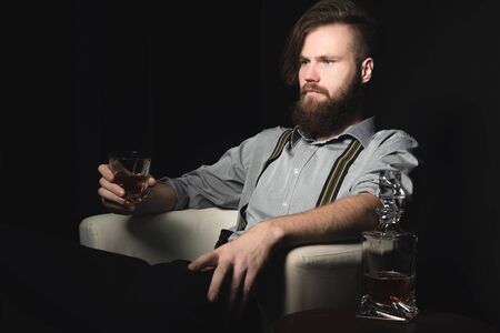 Portrait of a pensive handsome young man, with a glass of alcohol in his hands, on an isolated black background. The concept of pastime, rest and relaxationの写真素材