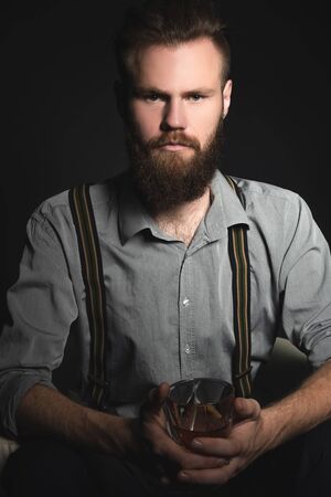 Portrait of a pensive handsome young man, with a glass of alcohol in his hands, on an isolated black background. The concept of pastime, rest and relaxationの写真素材