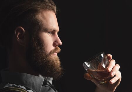 Portrait of a pensive handsome young man, with a glass of alcohol in his hands, on an isolated black background. The concept of pastime, rest and relaxationの写真素材