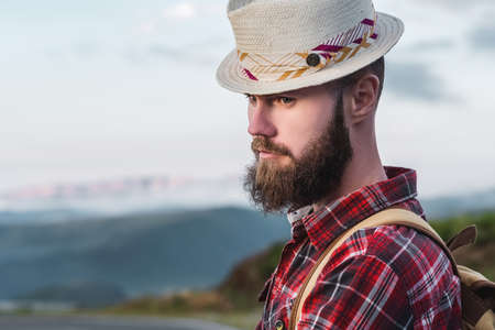 Handsome young man in a straw hat and a plaid shirt, stands, against the backdrop of beautiful mountains at sunset. The concept of freedom and hitchhikingの写真素材