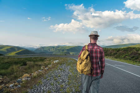 Handsome young man in a straw hat and a plaid shirt, walks along the road, against the backdrop of beautiful mountains at sunset. The concept of freedom and hitchhikingの写真素材