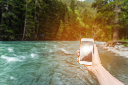 First-person view of a female hand with a phone on a background of a beautiful river and forest in...