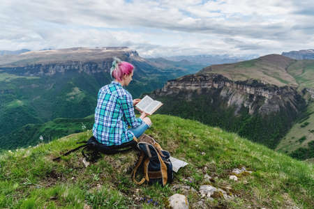 Beautiful young girl reads a book on a background of beautiful mountains. The concept of hanging out, relaxing and working.の写真素材