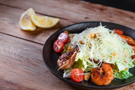 Appetizing Caesar salad with shrimps in a black plate on a wooden table next to a lemon. The concept...