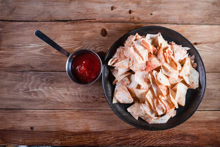 Potato chips in a plate next to ketchup on a wooden table. The concept of not healthy food, extra...