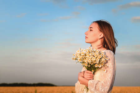Beautiful woman in a white dress with a bouquet of flowers in a wheat field at sunset. Free...
