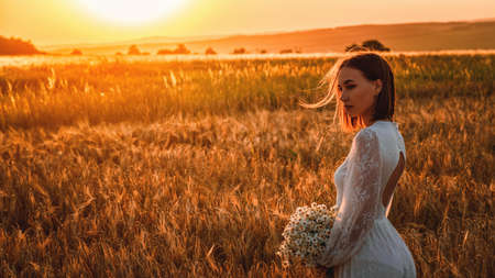 Beautiful woman in a white dress with a bouquet of flowers in a wheat field at sunset. Free lifestyle conceptの写真素材