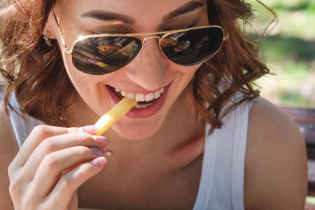 Beautiful, young woman eating fries in the street. The concept of fast food, food delivery and lunch in natureの写真素材