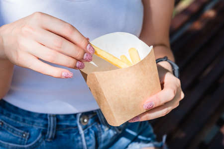 A slender girl holds a cardboard box with fried potatoes in her hands, close-up. Street food concept