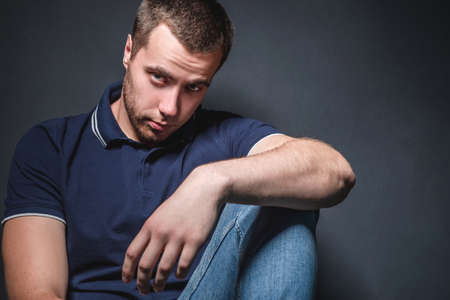 Young attractive man in blue jeans and a polo shirt sits on the floor near the wall in the studio. Youth and attractiveness conceptの写真素材