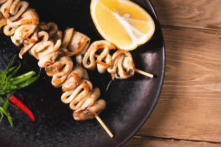 Fried squid bbq on skewers with lemon lies on a dark plate. Restaurant food and service concept