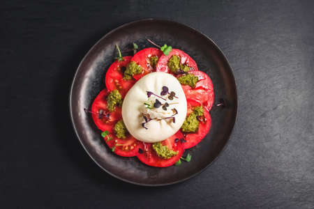 Salad of burrata cheese, tomatoes and pesto sauce in a black plate, on a dark background. Farm food and table setting conceptの写真素材
